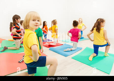 Vue de côté portrait de garçon enfant blonde, debout sur genou tenant la main sur sa taille, au cours de l'activité gymnastique en salle de remise en forme avec entraîneur féminin Banque D'Images