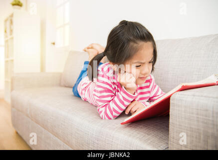 Little asian chinese girl couchée sur le canapé histoire lecture livre dur et difficile à comprendre le contenu dans le salon à la maison. fa Banque D'Images