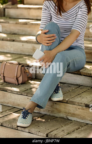 Jeune femme en bleu jeans et baskets à rayures se trouve sur d'anciennes marches de bois et est titulaire d'un téléphone dans ses mains. Focus sélectif. Banque D'Images