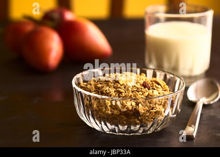 Céréales Petit déjeuner hors de flocons d'avoine, sésame, miel, amandes et les fruits secs (noix de coco, pomme, banane, raisins secs) dans un bol en verre avec un verre de lait et tamarillo dans le dos (Selective Focus, Focus sur les morceaux de fruits secs sur le dessus de la céréale) Banque D'Images