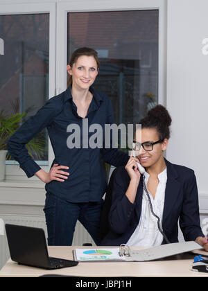 Eine junge Büroangestellte sitzt am Schreibtisch und vor einem Aktenordner telefoniert. Eine weitere Frau steht neben ihr und schaut zur Kamera. Banque D'Images