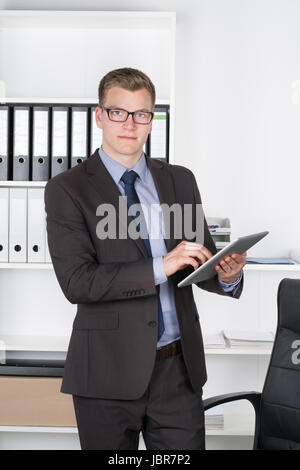 Junger Geschäftsmann mit Brille steht im Büro und vor einem Regal hält ein comprimé. Der Mann schaut zur Kamera. Banque D'Images