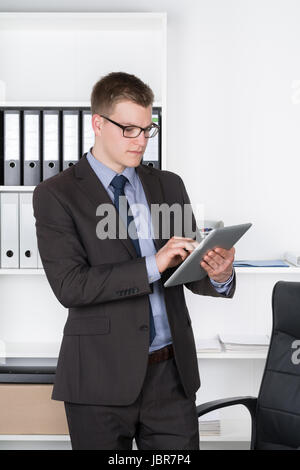 Junger Geschäftsmann mit Brille steht im Büro und vor einem Regal hält ein comprimé. Der Mann schaut auf das comprimé. Banque D'Images