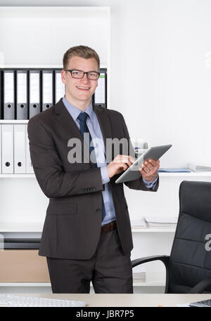 Junger Geschäftsmann mit Brille steht im Büro und vor einem Regal hält ein comprimé. Der Mann schaut zur Kamera und lächelt. Banque D'Images