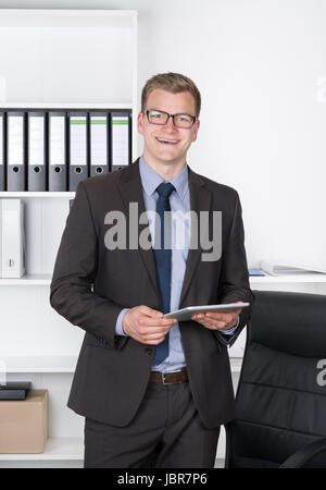 Junger Geschäftsmann mit Brille steht im Büro und vor einem Regal hält ein comprimé. Der Mann schaut zur Kamera und lächelt. Banque D'Images