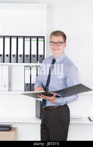 Junger Geschäftsmann mit Brille steht vor einem Regal und hält einen geöffneten Aktenorder. Der Mann schaut zur Kamera und lächelt. Banque D'Images