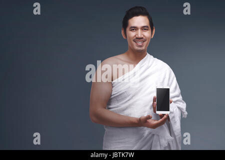 Portrait of asian musulman dans l'ihrâm dress holding smartphone sur fond sombre Banque D'Images