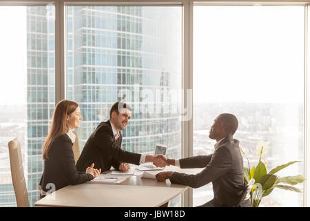 Friendly caucasian les employeurs et certains africains-américains au cours d'entrevue d'emploi requérant assis à un bureau avec grande grande fenêtre ur Banque D'Images