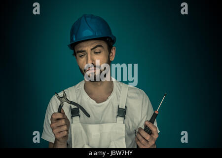 Jolie jeune barbu debout workman lorgne ses outils holding up une paire pf une pince et d'un tournevis comme il décide ce qui est bon d'utiliser Banque D'Images