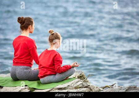 Vue arrière du jeune mère et fille faisant du yoga exercices sur la pierre près de la rivière. Belle Mère et fille dans le sportwear et pieds nus. Banque D'Images