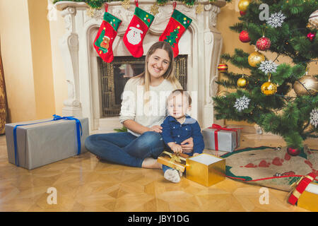 Heureux jeune mère assise avec son bébé sur le plancher à côté de cheminée de salon et l'arbre de Noël Banque D'Images