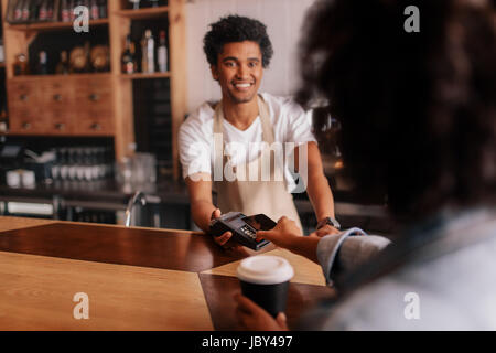 La clientèle féminine de vos paiements sur téléphone mobile au compteur dans cafe avec jeune homme. Barista holding credit card machine de lecture en face de femme Banque D'Images