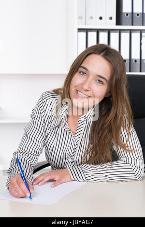 Eine junge, lächelnde schreibt Frau mit blauem Stift auf einem Blatt Papier am Schreibtisch Büro im. Im Hintergrund steht ein Regal. Die Frau schaut zur Kamera. Banque D'Images