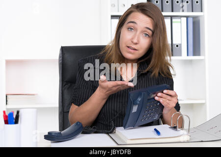Zeigt eine junge Frau mit einer skeptisch Hand auf ein Telefon, welches sie in der anderen part während der sie am Schreibtisch Büro im sitzt. Im Hintergrund steht ein Regal. Die Frau schaut zur Kamera.