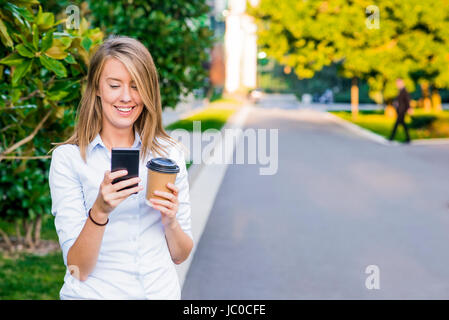 Portrait d'une femme joyeuse en utilisant son smartphone et l'exécution des dossiers de travail, debout contre un ciel bleu ensoleillé. Banque D'Images