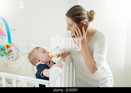 Portrait de jeune mère parler par téléphone et nourrir son bébé de la bouteille Banque D'Images