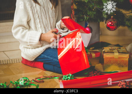 Libre de droit de la fille assise sous l'arbre de Noël et de coupe papier rouge avec des ciseaux pour la décoration présente Banque D'Images