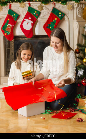 Beautiful smiling girl et mère wrapping Christmas gifts en papier rouge et attacher avec du ruban d'or Banque D'Images