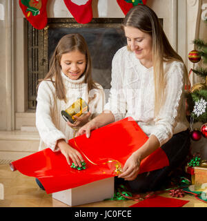 Portrait of happy young mère et fille cute cadeaux de Noël d'emballage en papier rouge et attacher avec du ruban d'or Banque D'Images