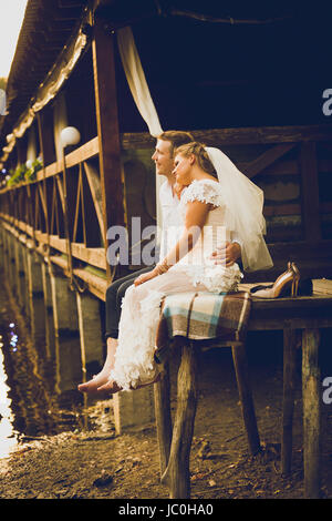 Photo de tons nouveaux mariés Mariés assis sur l'ancienne jetée et à la recherche au coucher du soleil Banque D'Images