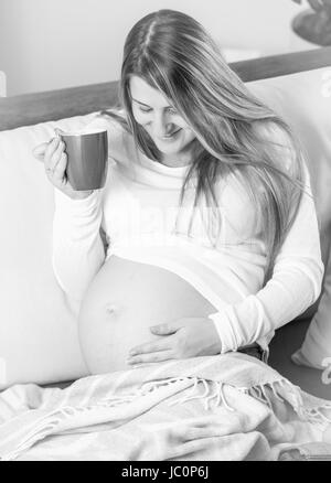 Photo en noir et blanc de smiling pregnant woman lying on sofa et boire le thé Banque D'Images