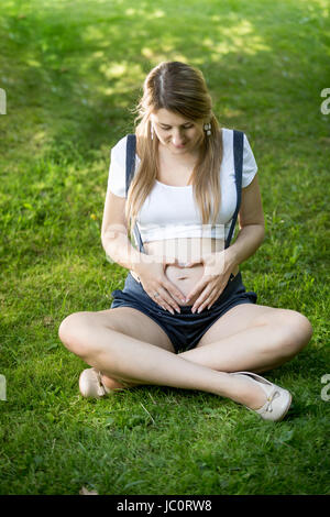 Jeune femme enceinte se détendre sur l'herbe et tenir la main à forme de coeur sur l'abdomen Banque D'Images