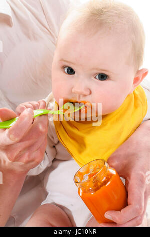 Erwachsene Frau sitzt auf einem weissen Stuhl und das haelt 6 Monate alte und im fuettert Bébé bras mit einem gelben Plastikloeffel Karottenbrei isoliert auf orangen, vor weissem Hintergrund. Banque D'Images