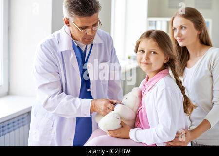 Smiling girl looking at camera tout en examinant son médecin dans les cliniques de l'ours en peluche Banque D'Images