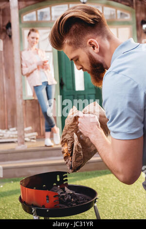 Jeune femme debout sur le porche et à la recherche à l'homme barbu tenant le charbon pour barbecue Banque D'Images