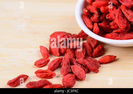 Le Goji séché berris renversé sur une planche à découper en bambou avec plus de goji dans une soucoupe blanche. Certains dans la soucoupe et quelques réparties sur le bambou. Banque D'Images