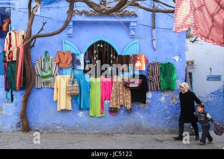 Une fille marche son frère à la maison de l'école au-delà d'un affichage des vêtements colorés, compensée par la suppression des murs bleus à Chefchaouen, Maroc, dont la médina (vieille ville) est célèbre pour ses bâtiments bleus. Banque D'Images