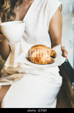 Petit-déjeuner italien ou français. Jeune femme blonde en robe blanche tenant des croissants frais et une tasse de cappuccino au café Banque D'Images