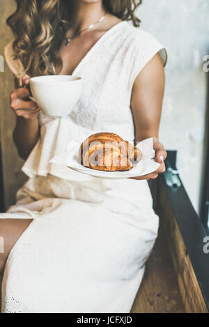 Petit-déjeuner italien ou français. Young blonde woman in white dress holding croissant frais dans la plaque et une tasse de cappuccino au café Banque D'Images