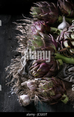 Tout crus artichauts violets humides organiques avec de l'ail sur les textiles sacs sur fond de bois sombre. Style rustique. Banque D'Images