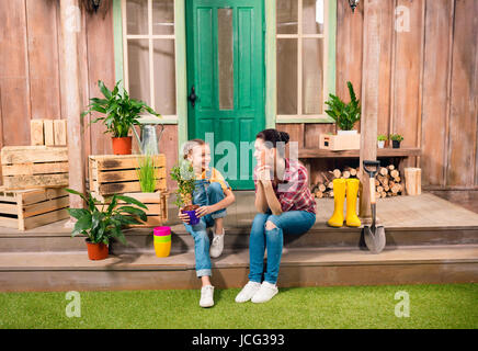 Happy mother and daughter with potted plant, assis sur le porche et souriant les uns les autres Banque D'Images