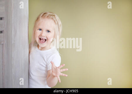 La petite fille blonde aux cheveux longs dans un t-short.La fille en peek de derrière la porte et a l'intention de faire peur à quelqu'un. Banque D'Images