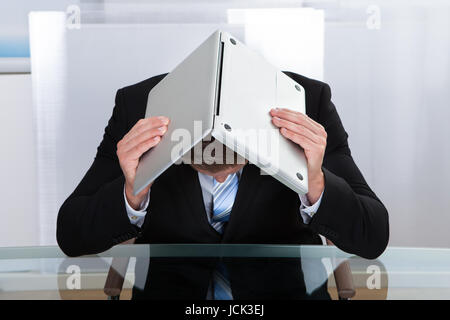 Déprimé businessman sitting at a table réfléchissant cachait sous son ordinateur portable comme il le maintient ouvert sur sa tête s'inclina Banque D'Images