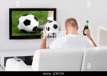 Heureux fan de foot un match à la télévision tenant un ballon au-dessus de sa tête comme il est assis sur un canapé confortable dans son salon Banque D'Images