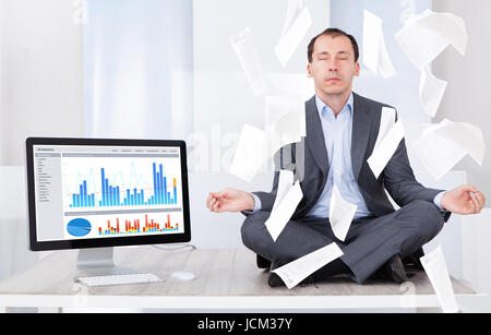 Toute la longueur de Mid adult businessman meditating tandis que documents sur lui par computer in office Banque D'Images