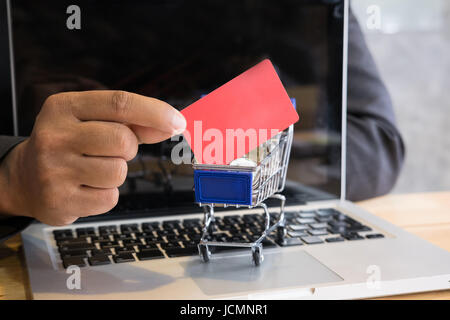 Jeune homme montrant une carte de crédit dans un mini supermarché panier chariot plein de pièces sur un ordinateur portable pour faire des achats en ligne. businessman acheter des biens fro Banque D'Images