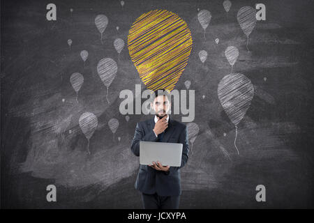 Thoughtful businessman holding laptop in front of blackboard chalk avec icônes de ballon Banque D'Images