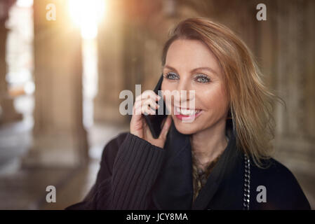 Jolie femme en conversation sur un smartphone à l'intérieur d'un grand bâtiment en souriant avec plaisir qu'elle écoute la conversation Banque D'Images