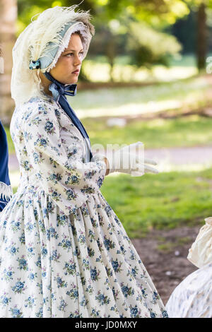 Pretty young woman, 14-15, woman standing in park habillés en costume victorien. Robe blanche à fleurs bleues et lacy hat attaché avec grand ruban bleu. Vue de côté. Banque D'Images