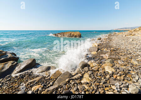 Les graviers de vagues se brisant sur l'eau, les cailloux et les rochers d'une plage vide dans la rude côte rocheuse de la Ligurie, au nord de l'Italie. Ciel bleu clair, large ang Banque D'Images