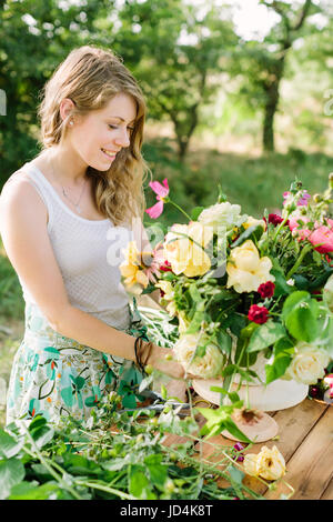 L'été, beauté, mode de vie, les arts et l'artisanat, concept de mariage - charmante jeune femme aux longs cheveux blonds se tenait à côté de la table de jardin en bois et de superbes bouquet composé de roses et de pivoines Banque D'Images