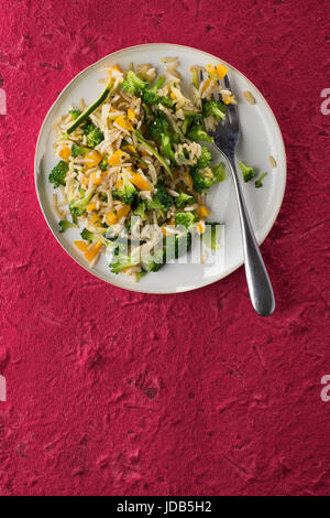 Un plat de riz frit vegan faite avec du riz basmati, le brocoli, la courgette et le poivron jaune, avec une fourchette, sur un fond texturé rouge vif. Banque D'Images