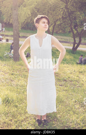 Jeune femme aux cheveux courts portant robe de lin blanc debout dans le parc, retour du soleil Banque D'Images
