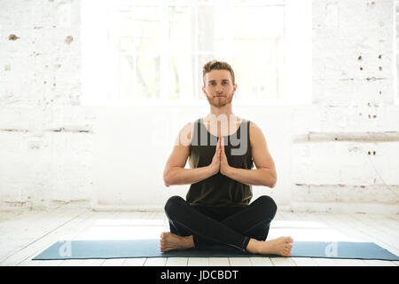 Young smiling guy assis sur un tapis de fitness et méditer dans la salle de sport Banque D'Images