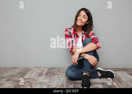 Photo de smiling young asian woman isolated over grey wall assis. En regardant la caméra. Banque D'Images