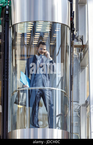 Businessman dans l'ascenseur en verre moderne de parler par le téléphone et la tenue de documents Banque D'Images